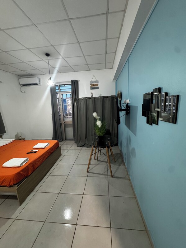 A hotel room with a bed, a small table with a plant, a wall-mounted air conditioner, and a window with curtains.