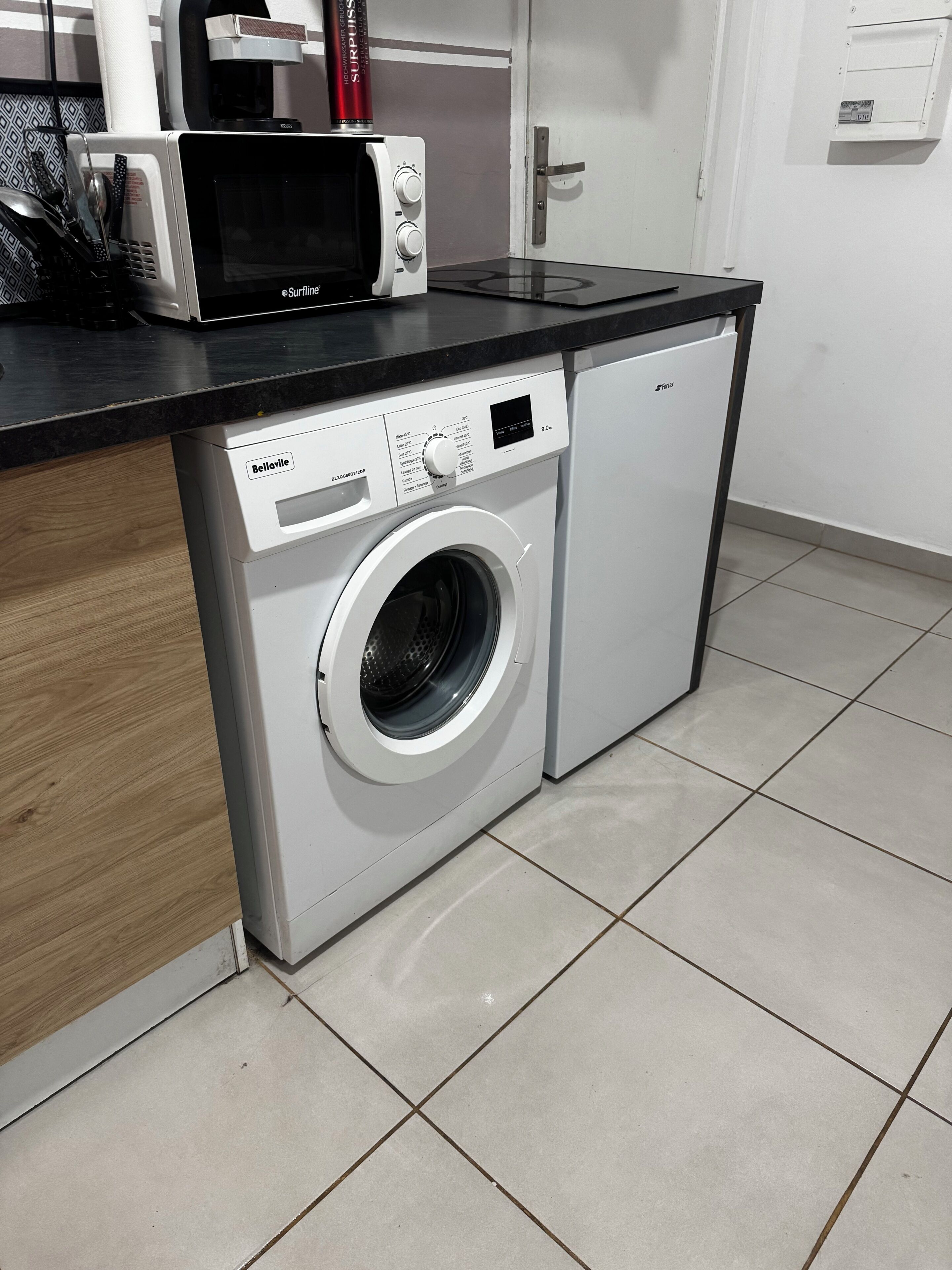 A compact washer and dryer unit with a control panel, placed on a tiled floor with a wooden cabinet adjacent to it.