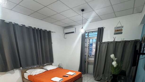 Ein Schlafzimmer mit Bett, Vorhängen, Klimaanlage, einem kleinen Tisch mit Pflanze und Blick durch ein Fenster.