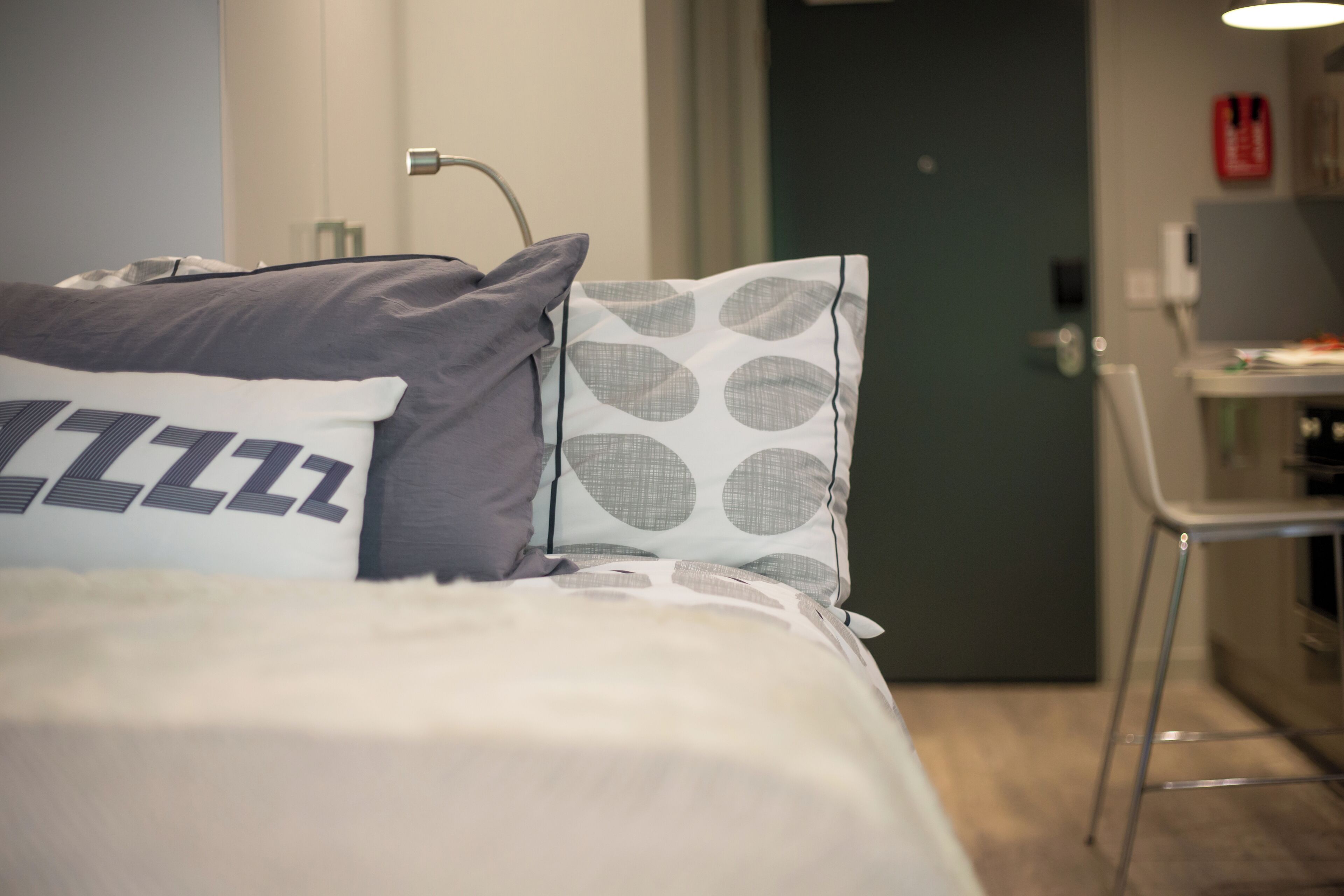 En snyggt bäddad säng med en prydlig kudde med ett iögonfallande mönster, ett sängbord med en lampa och en dörr som leder in till ett köksutrymme.