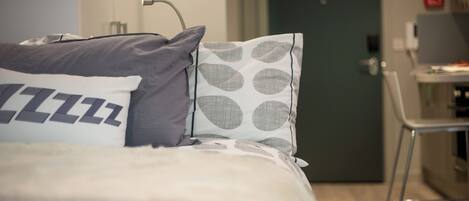 En snyggt bäddad säng med en prydlig kudde med ett iögonfallande mönster, ett sängbord med en lampa och en dörr som leder in till ett köksutrymme.