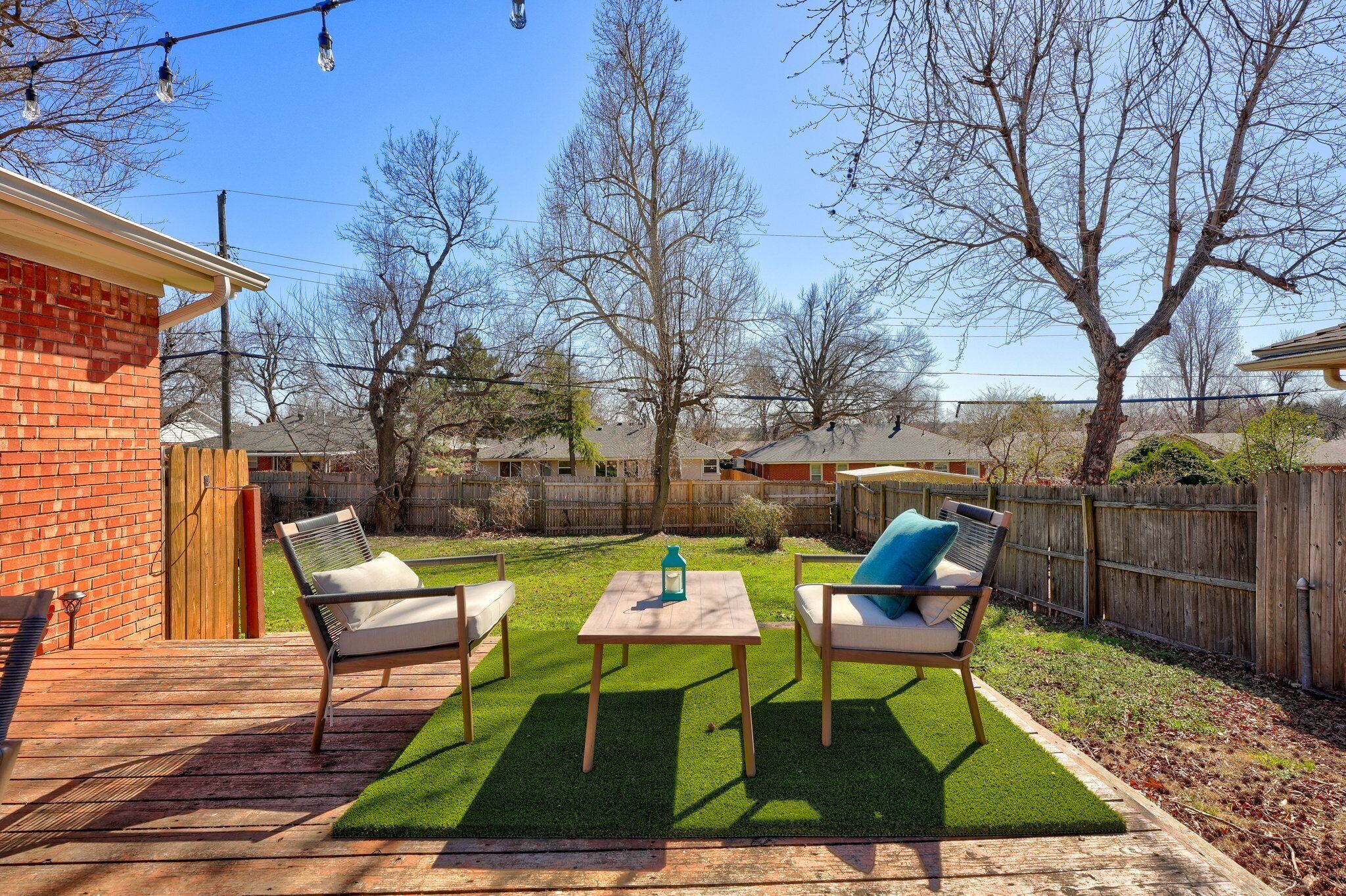 A backyard with a wooden deck, two lawn chairs, a small table, and a green artificial turf.