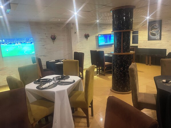 A restaurant with tables set for dining, a large pillar, and a TV screen displaying a sports event. - Maracanaluxury Hotel & Suites (Ibadan)