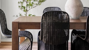Un comedor con una mesa de madera, sillas de ratán negro, un florero blanco con flores y un panel de pared tejido.