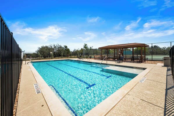 A rectangular swimming pool with clear blue water, surrounded by a tiled deck and a covered seating area.