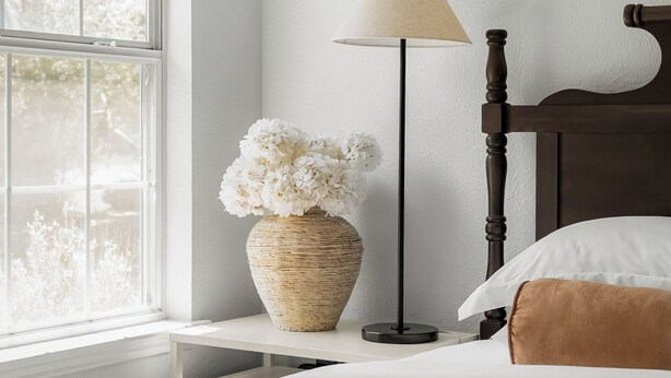 Una cama bien hecha con sábanas blancas, una lámpara de noche, un florero con flores blancas y una ventana con vistas a árboles.