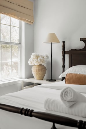 A neatly made bed with white linens, a bedside lamp, a vase with white flowers, and a window with a view of trees.