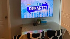 A living room with a flat-screen TV mounted on the wall displaying 'DISKARTE!' on a cityscape background. There are four chairs with black and white patterns around a small table. - Azure Urban Resort Residences (Paranaque)