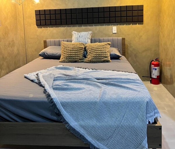 A bed with a grey headboard, two pillows, a blue blanket, and a fire extinguisher on the wall.