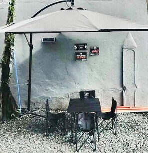Un área de estar al aire libre con una mesa y sillas bajo un gran paraguas, una unidad de aire acondicionado instalada en la pared y dos letreros que solicitan silencio y prohíben fumar.