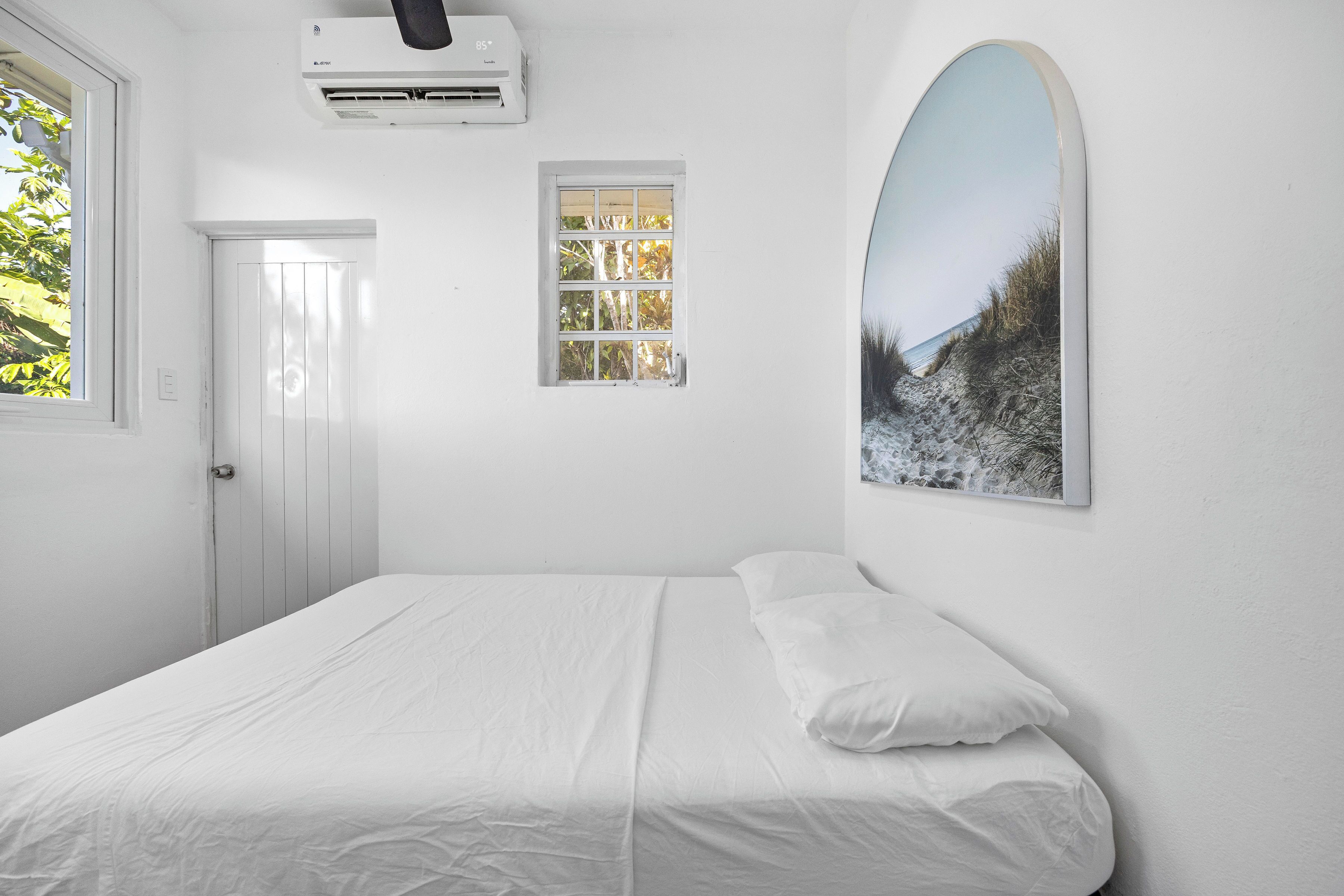 A bedroom with a bed, a window, an air conditioning unit, and a framed picture on the wall.