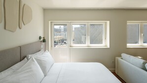 Une chambre moderne avec un grand lit, un canapé et deux fenêtres donnant sur la ville.
