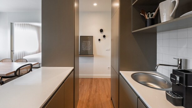 A modern kitchen with a built-in oven, a sink, and a coffee machine. There is a dining area with a table and chairs visible in the background.