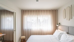 A hotel room with a large bed, a desk, a chair, and a small stool.