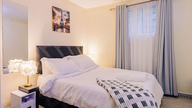 Un lit bien fait, avec du linge de lit blanc, une tête de lit noire et un plaid à motifs. On y voit une petite table de chevet avec une lampe et un objet décoratif. Un tableau représentant la Tour Eiffel est accroché au mur.