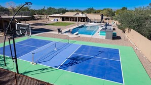 Pista de tenis con piscina y zona de descanso, rodeada por una valla y árboles.
