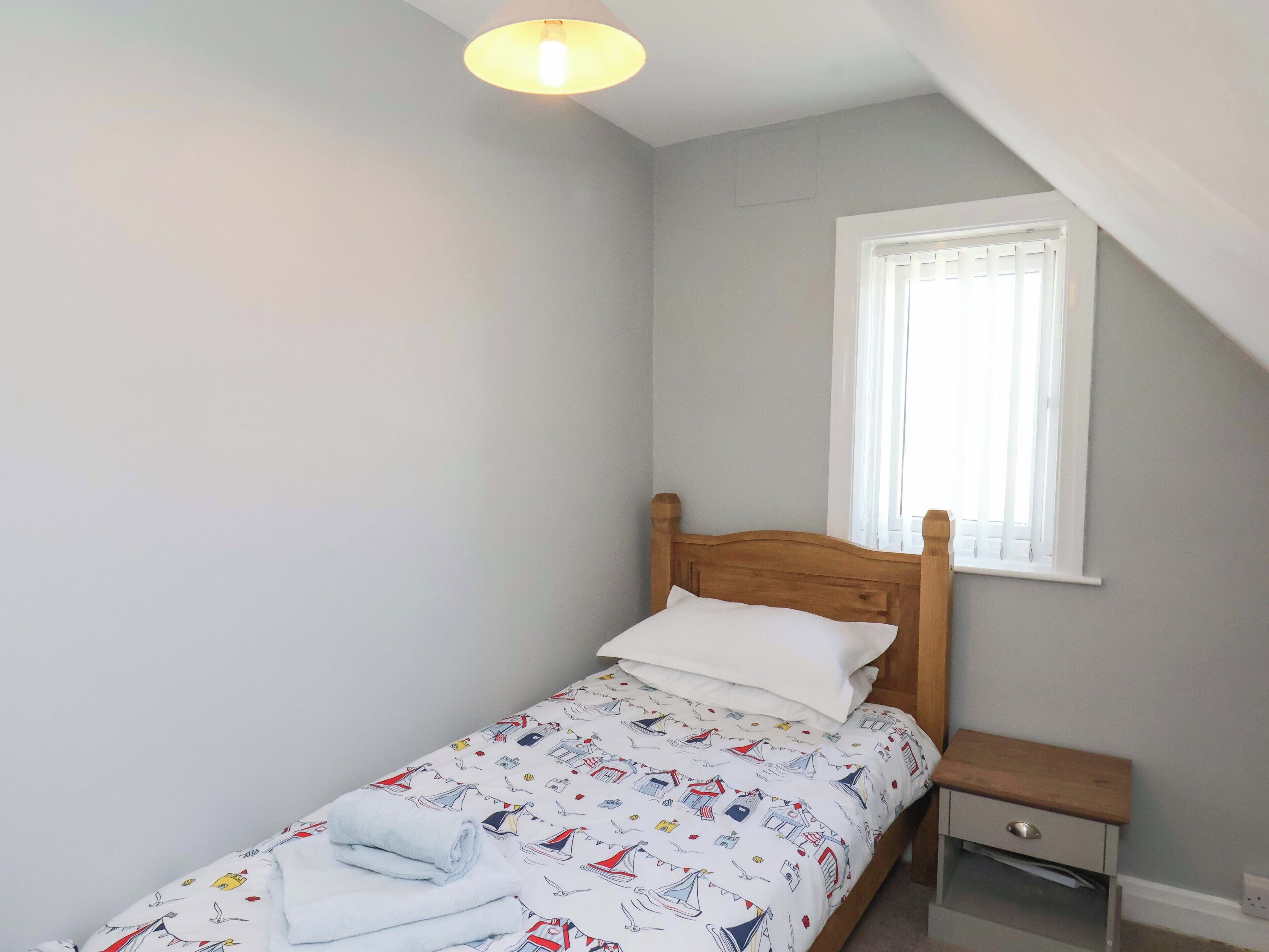 A small, neatly made bed with a wooden headboard in a room with a sloped ceiling and a window with white blinds.