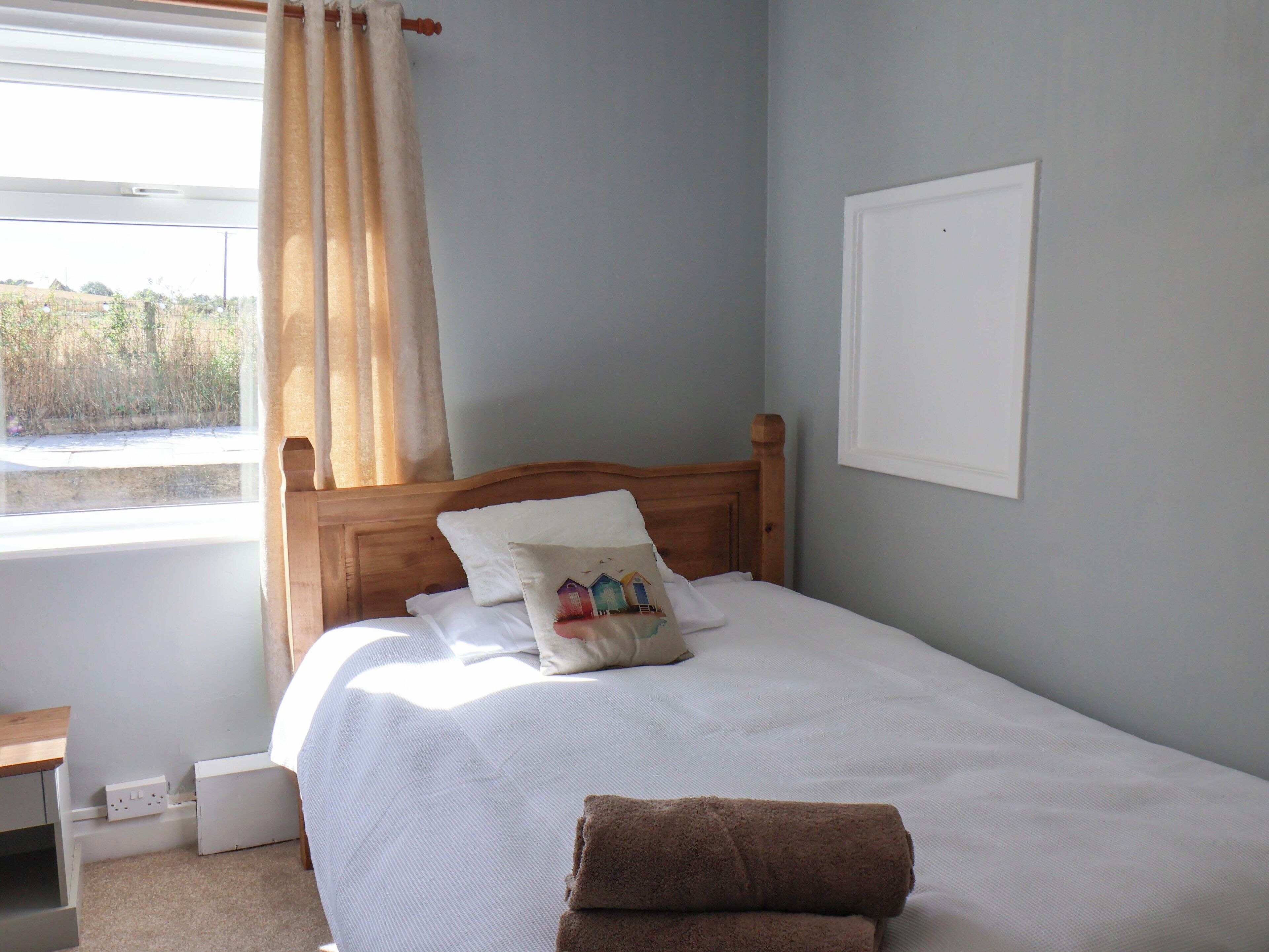 Una cama bien hecha con cabecera de madera, una ventana con cortinas y vistas al exterior con vegetación.