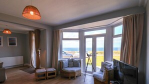 Ein geräumiges Wohnzimmer mit großem Fenster und Meerblick, mit Korbmöbeln, einem Fernseher und einem Teleskop.