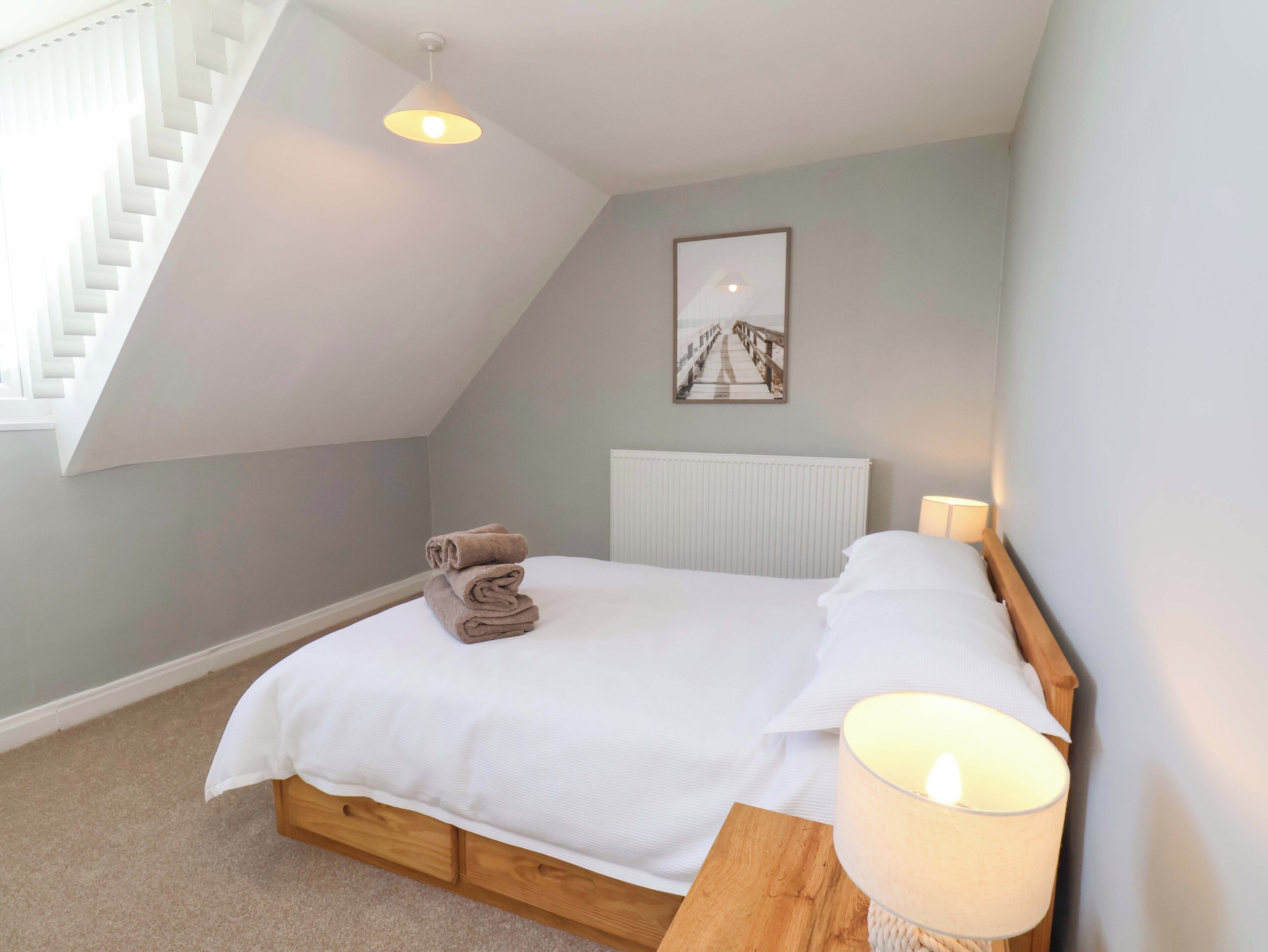 Un dormitorio con techo inclinado, una cama con ropa blanca, una toalla enrollada sobre la cama, una mesita de noche con una lámpara y un cuadro en la pared.