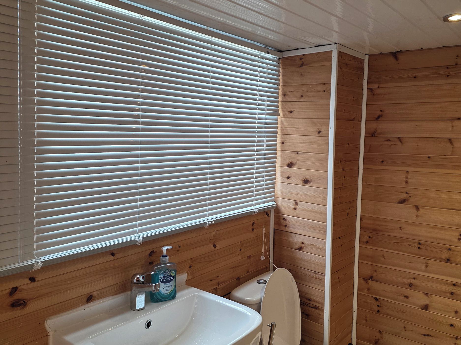 Ein Badezimmer mit Holzeinrichtung, einem Waschbecken, einer Toilette und einem Fenster mit Jalousien.