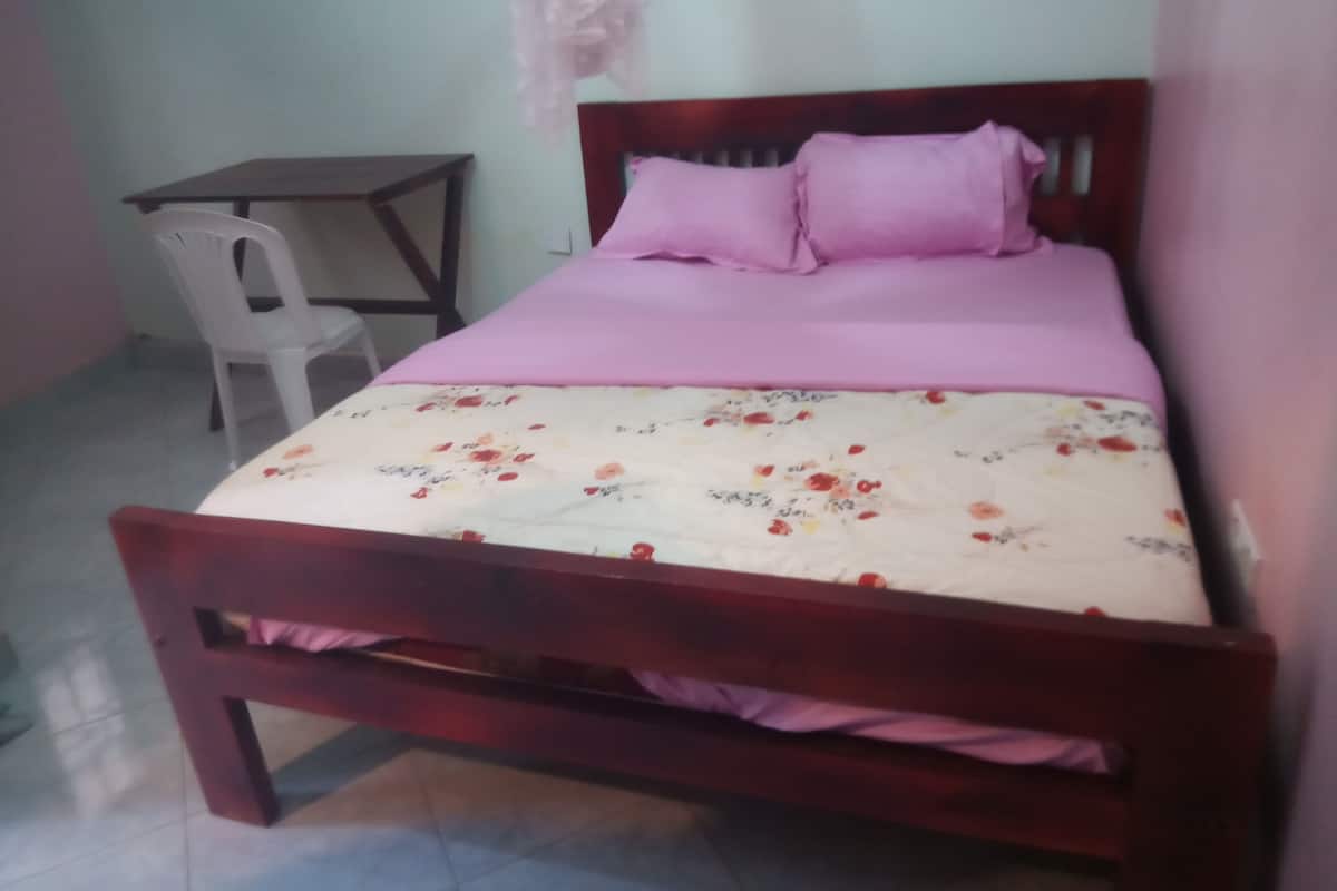 Ein Einzelbett mit Holzrahmen, einer blumigen Tagesdecke und zwei rosafarbenen Kissen. Eine kleine Holztisch und ein weißer Plastikstuhl befinden sich links vom Bett.