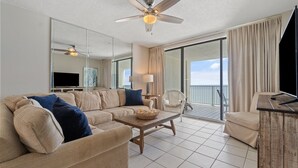 A spacious living room with a large sofa, a coffee table, and a TV. There is a balcony with a view of the ocean. - Pelican Pointe 1203 (Orange Beach)
