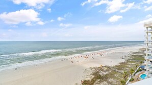 A beach with umbrellas and people, a building with balconies, and a clear sky. - Pelican Pointe 1203 (Orange Beach)