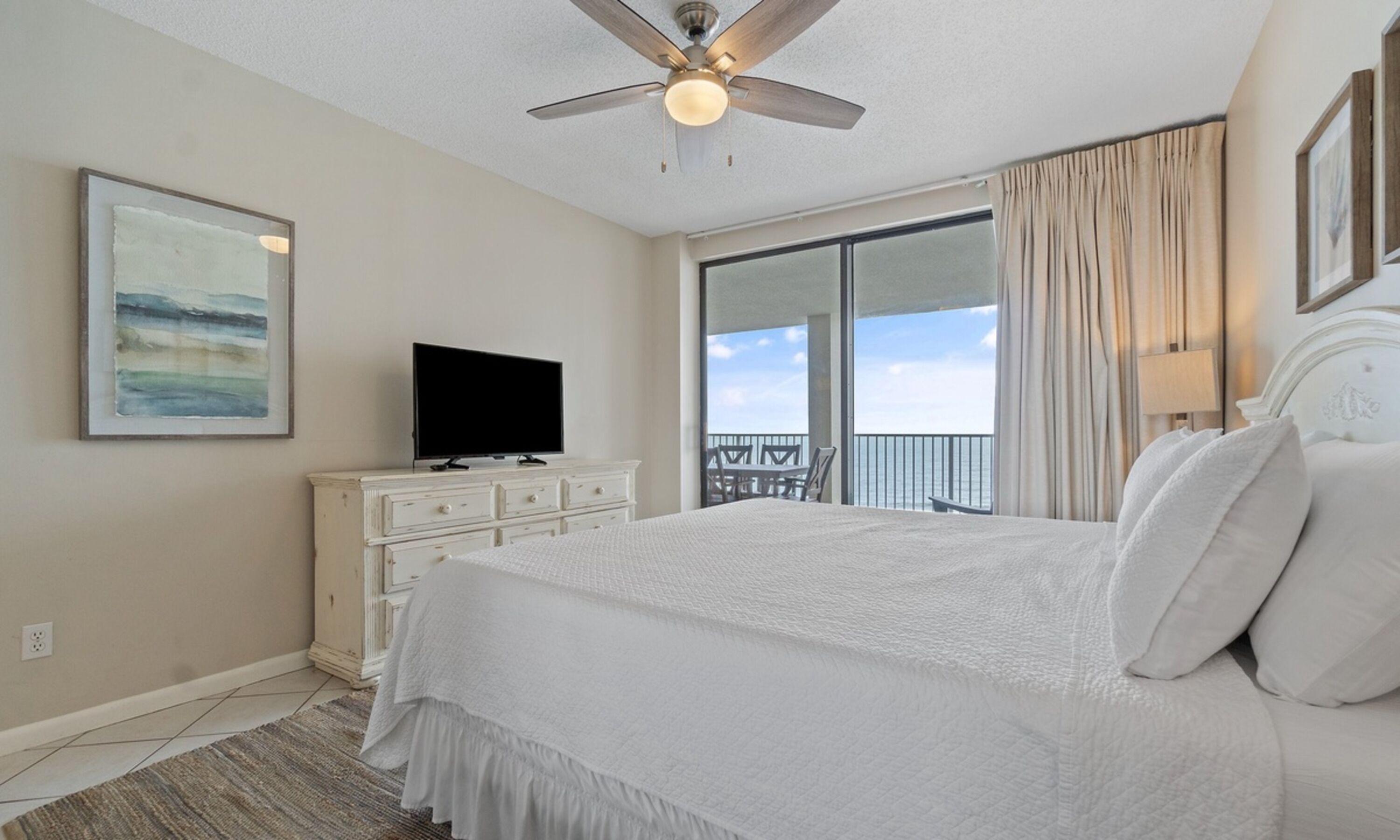 A hotel room with a large bed, a ceiling fan, a television on a dresser, a balcony with a view, and a painting on the wall.