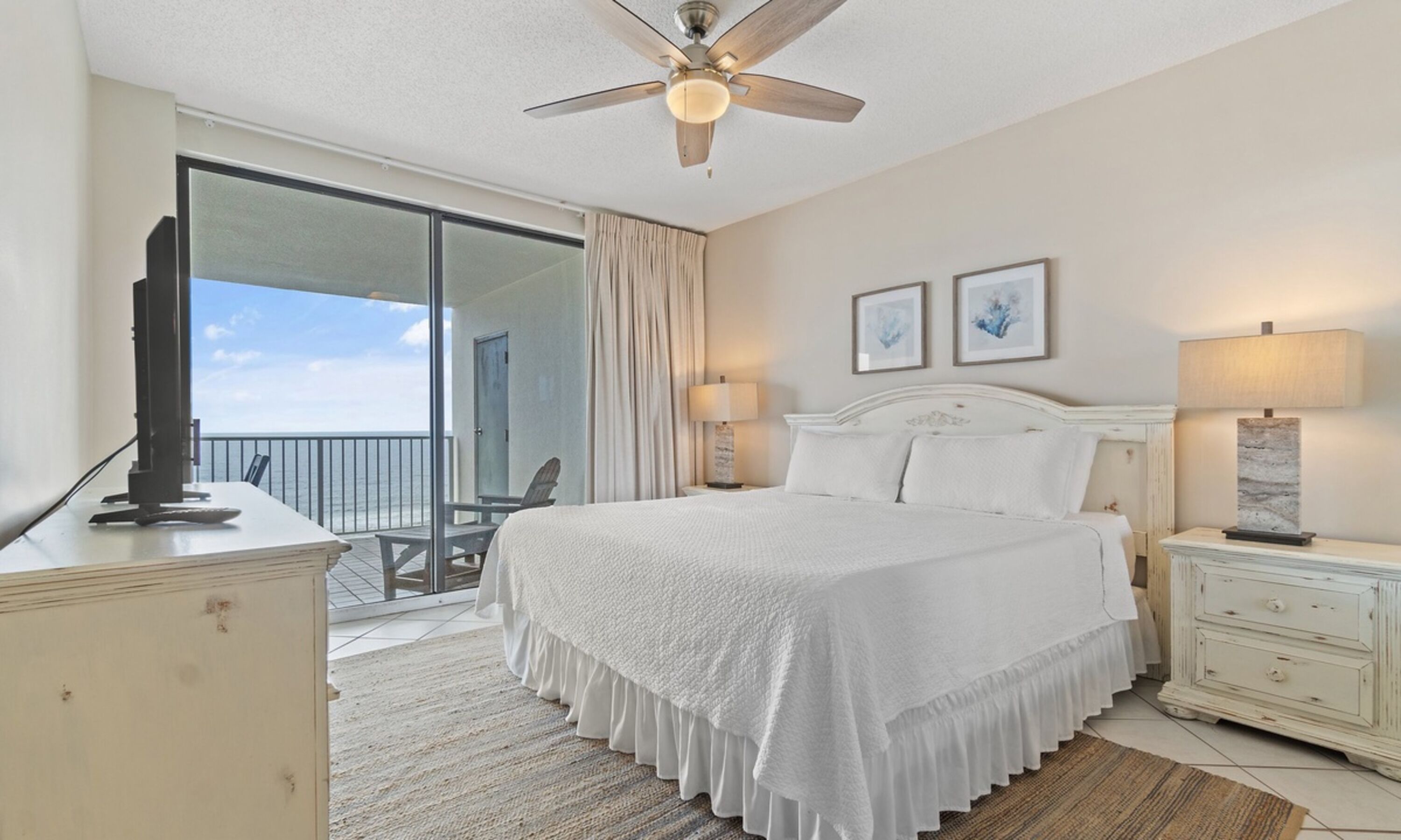 A bedroom with a large bed, a ceiling fan, a desk with a computer, a chair, and a view of the ocean through a sliding door.
