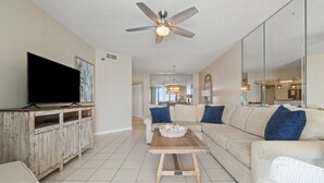 A living room with a flat-screen TV, a wooden cabinet, a sofa with blue cushions, a coffee table, and a ceiling fan. - Pelican Pointe 1203 (Orange Beach)