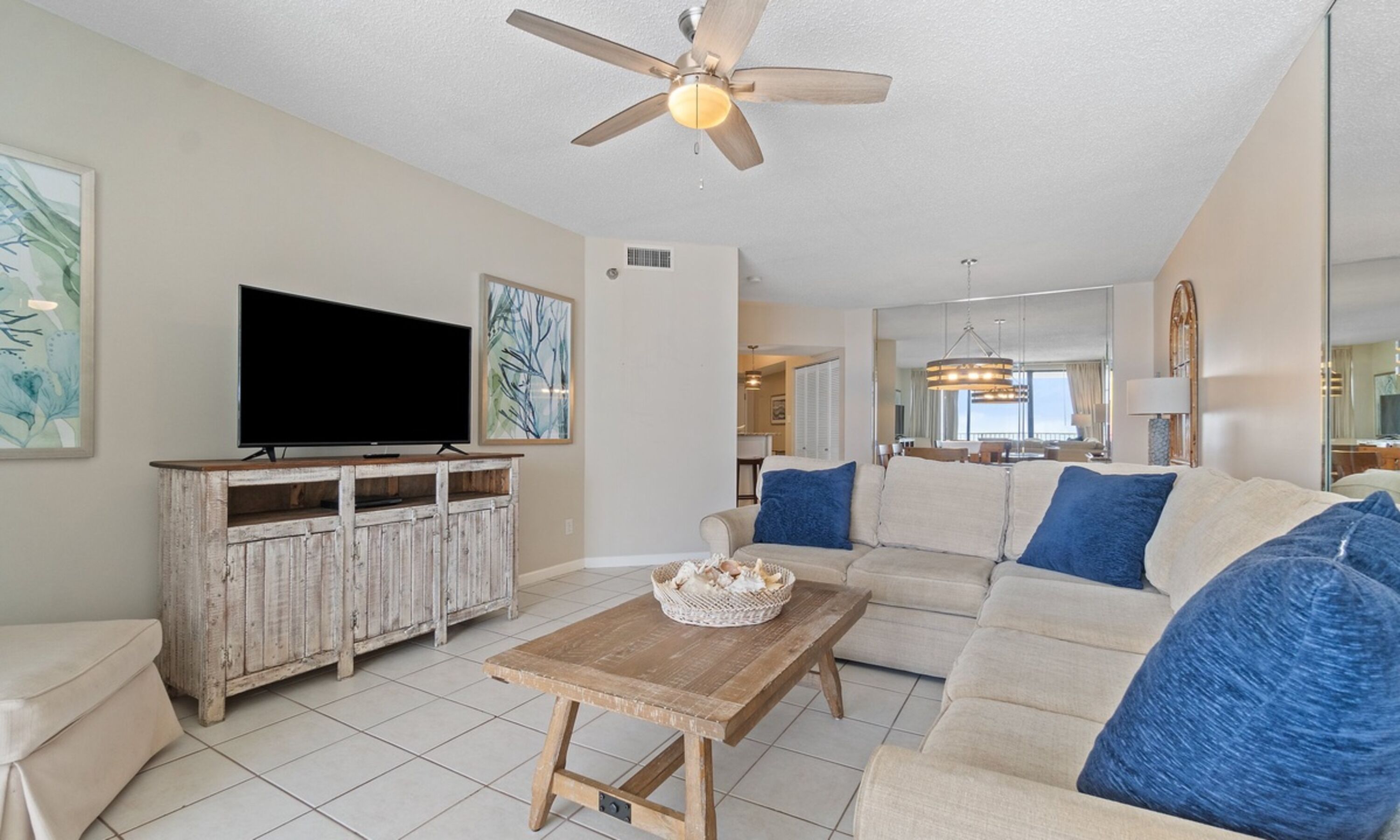 A living room with a flat-screen TV, a wooden coffee table, a beige sofa with blue cushions, and a ceiling fan.