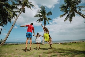 A family of four with a man in a red shirt and blue shorts, a woman in a yellow dress, and two children, one in a white dress, standing on a grassy area with palm trees and a view of the ocean.