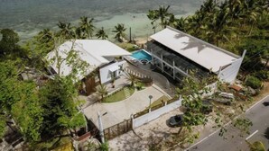 A modern two-story house with a swimming pool, surrounded by tropical vegetation and a beach.