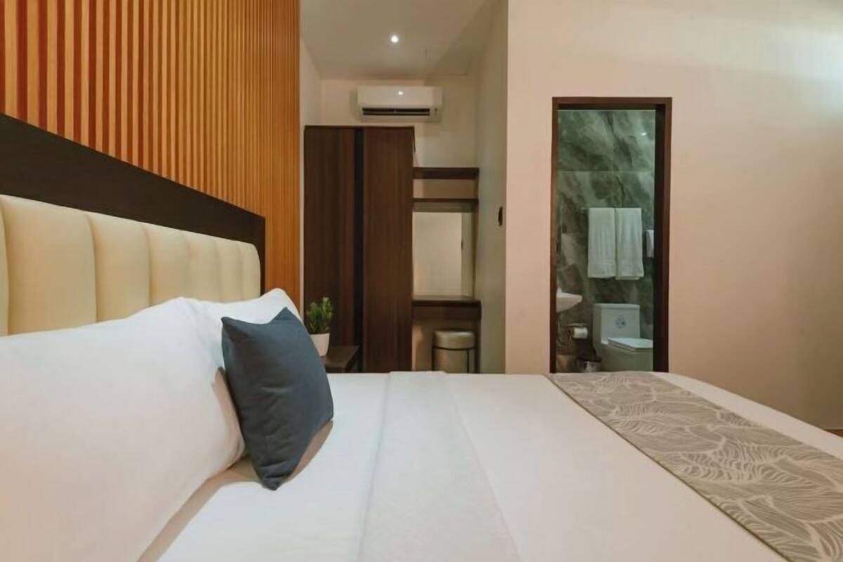 A hotel room with a large bed, a wooden headboard, a small table with a plant, and a bathroom visible through an open door.