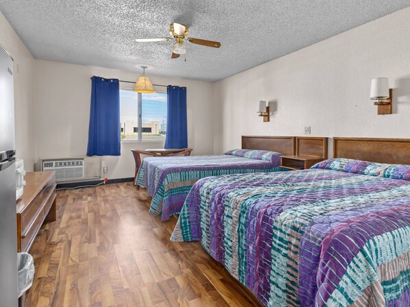 A hotel room with two beds, a ceiling fan, a window with curtains, a wooden desk, and a heater.