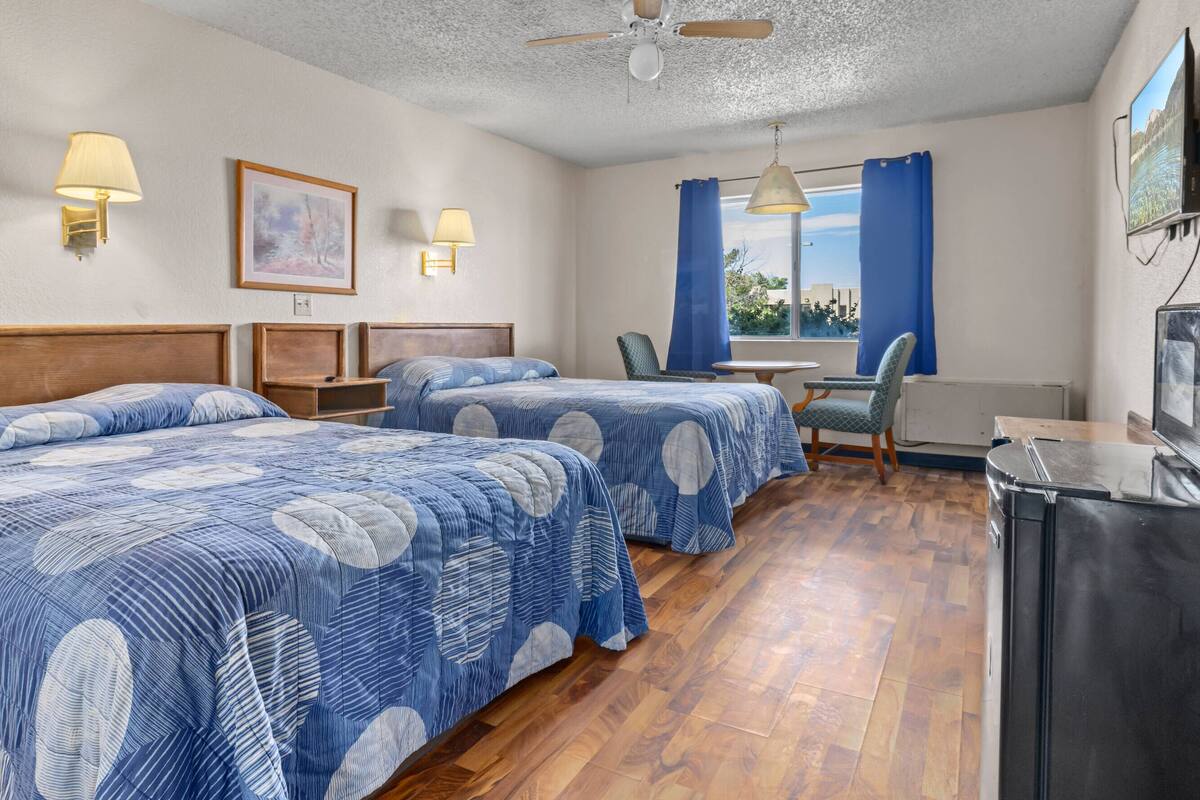 A hotel room with two beds, a desk, a chair, a ceiling fan, and a TV.