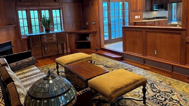 A spacious living room with a large window, wooden furniture, and a view into the kitchen.