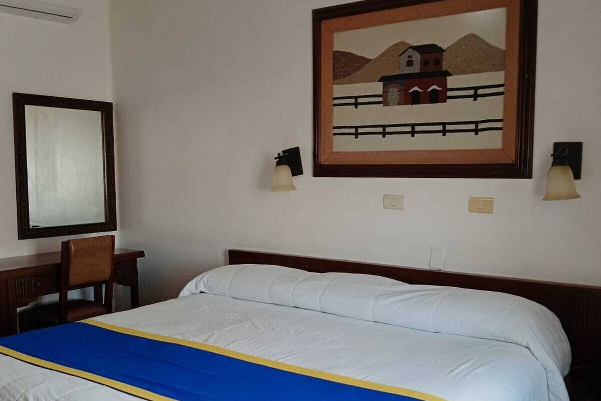 A hotel room with a large bed, a desk, and a chair. There is a painting on the wall and two wall-mounted lamps.