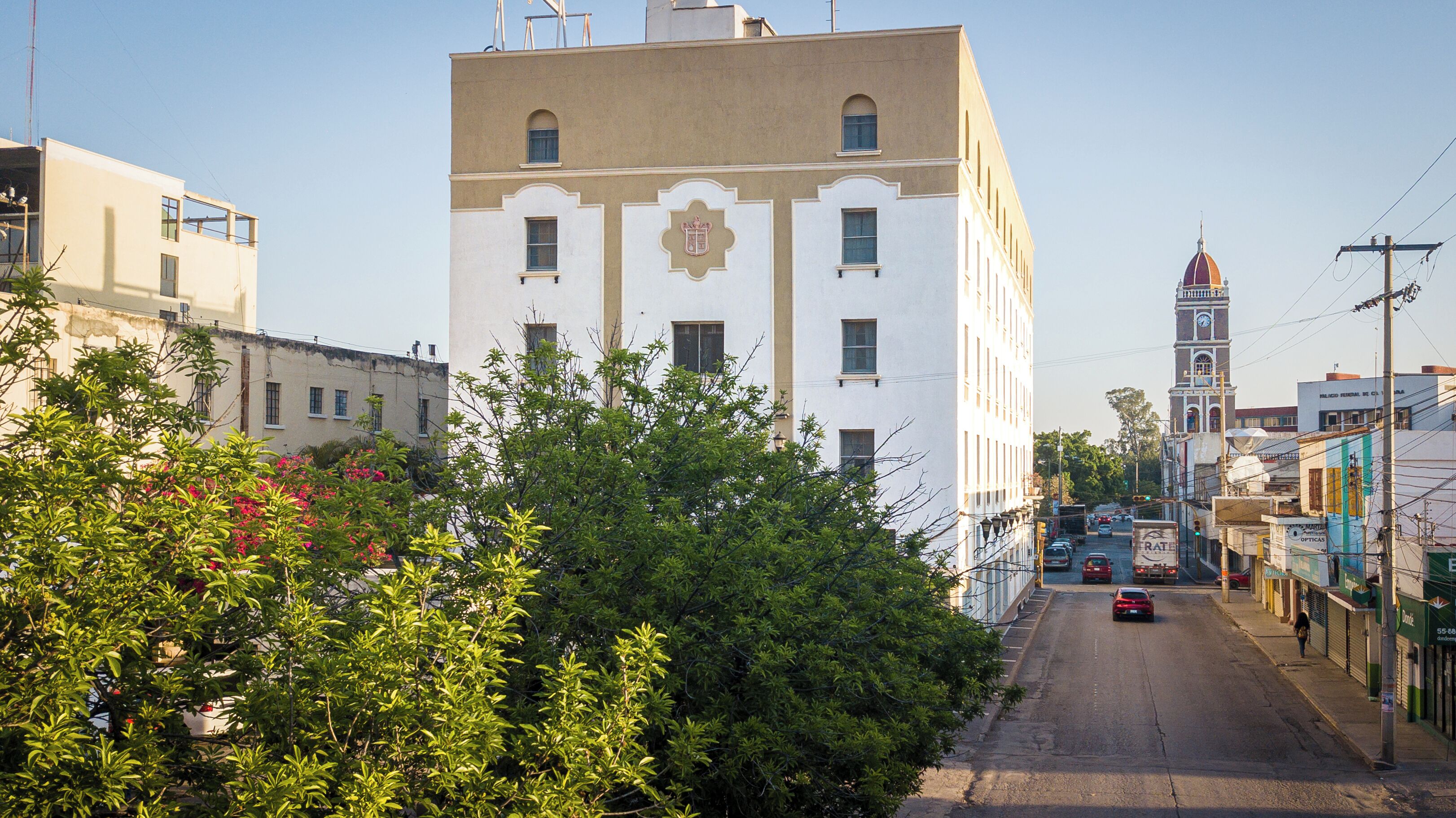 A street view with a prominent white building with a clock tower, surrounded by greenery and other buildings.