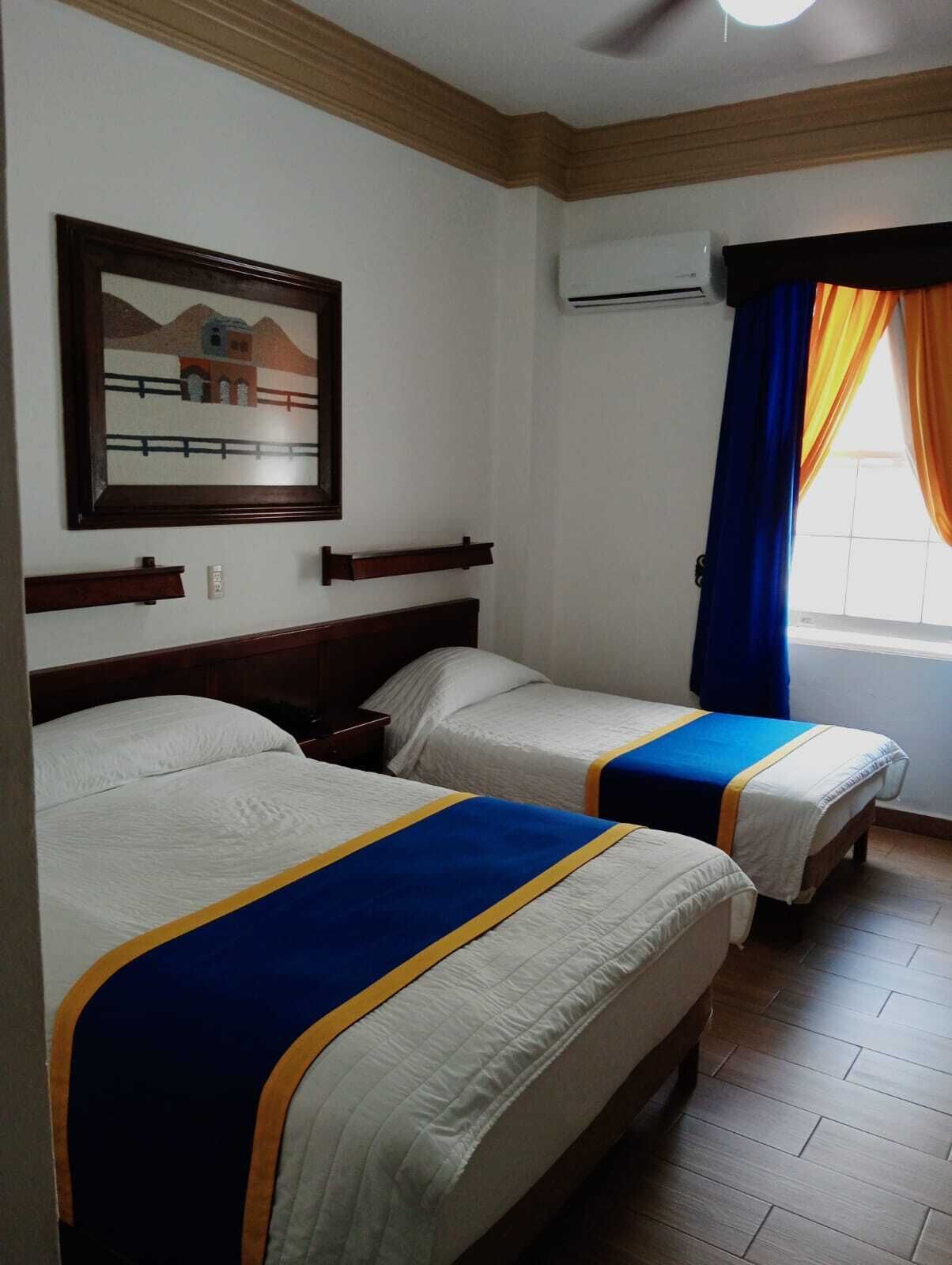 A hotel room with two beds, a painting on the wall, an air conditioning unit, and a window with curtains.