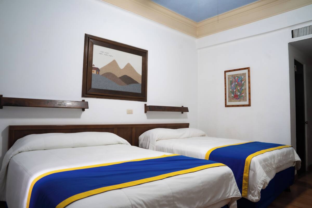 A hotel room with two beds, each with a blue and yellow bedspread, a wooden headboard, and a framed picture on the wall.