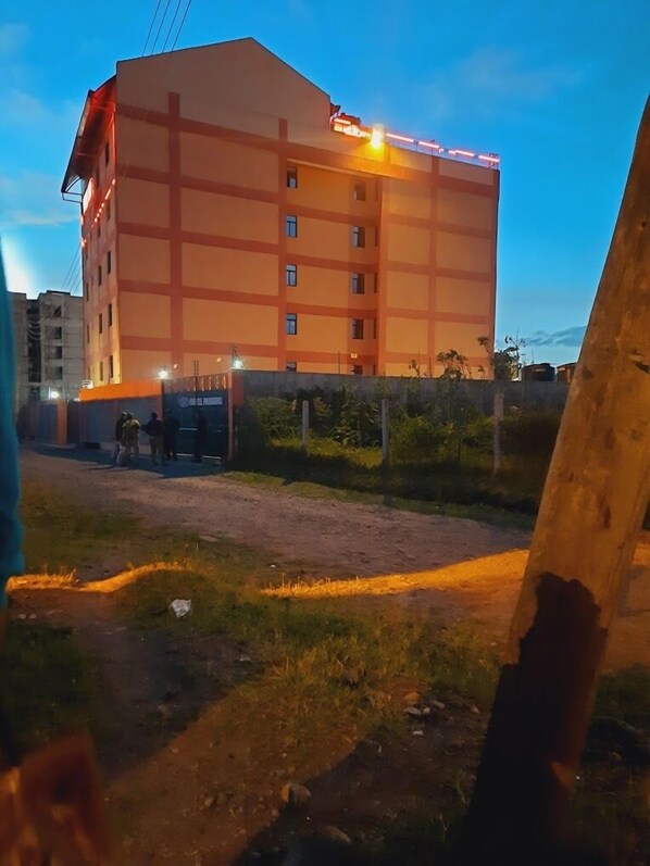 Ein mehrstöckiges Gebäude mit flachem Dach und einer Reihe von Fenstern. Vor dem Gebäude befindet sich ein gepflasterter Bereich mit Grünpflanzen und einem Zaun.