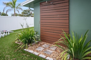 A modern building with a brown exterior wall, a small garden area with plants and stepping stones, and a white fence in the background.