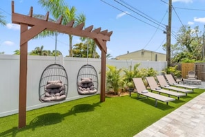 A backyard with artificial grass, a pergola with hanging egg chairs, and a row of sun loungers.