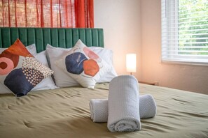 A neatly made bed with a green headboard, multiple pillows, and a rolled towel on a beige bedspread.