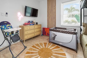 Ein Spielbereich für Kinder mit Hochstuhl, einer Kommode, einem Babybett, einem Fernseher und einem Fenster mit Jalousien.