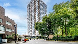 A modern high-rise building with a corner design, surrounded by trees and a street. - Stylish 2BR Flat | Oxford Street Locale (Greater London)