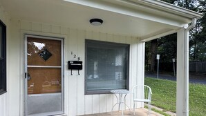 Una casa con puerta principal, buzón, ventana con persianas, una pequeña mesa y silla, y felpudo de bienvenida.