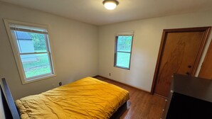 Una habitación pequeña con cama individual, colcha amarilla, dos ventanas, una puerta y una lámpara en el techo.